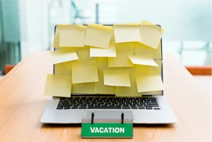 A laptop covered in multiple yellow sticky notes, sitting on a desk, with a green sign in front that reads "VACATION" symbolizing an overwhelming workload left behind while someone is out of office.